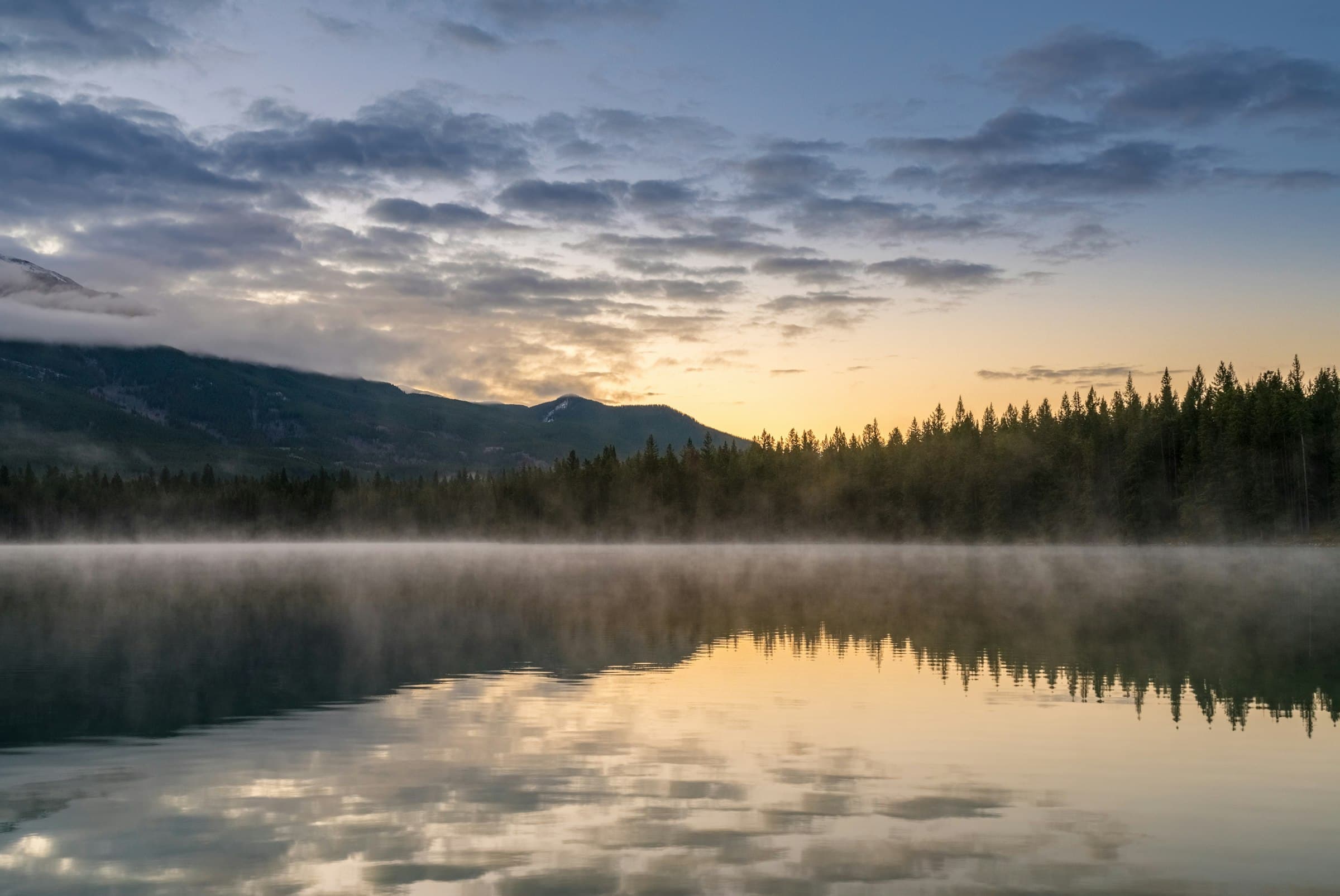 Calm still body of water at dawn reflecting a soft gold and grey sky.